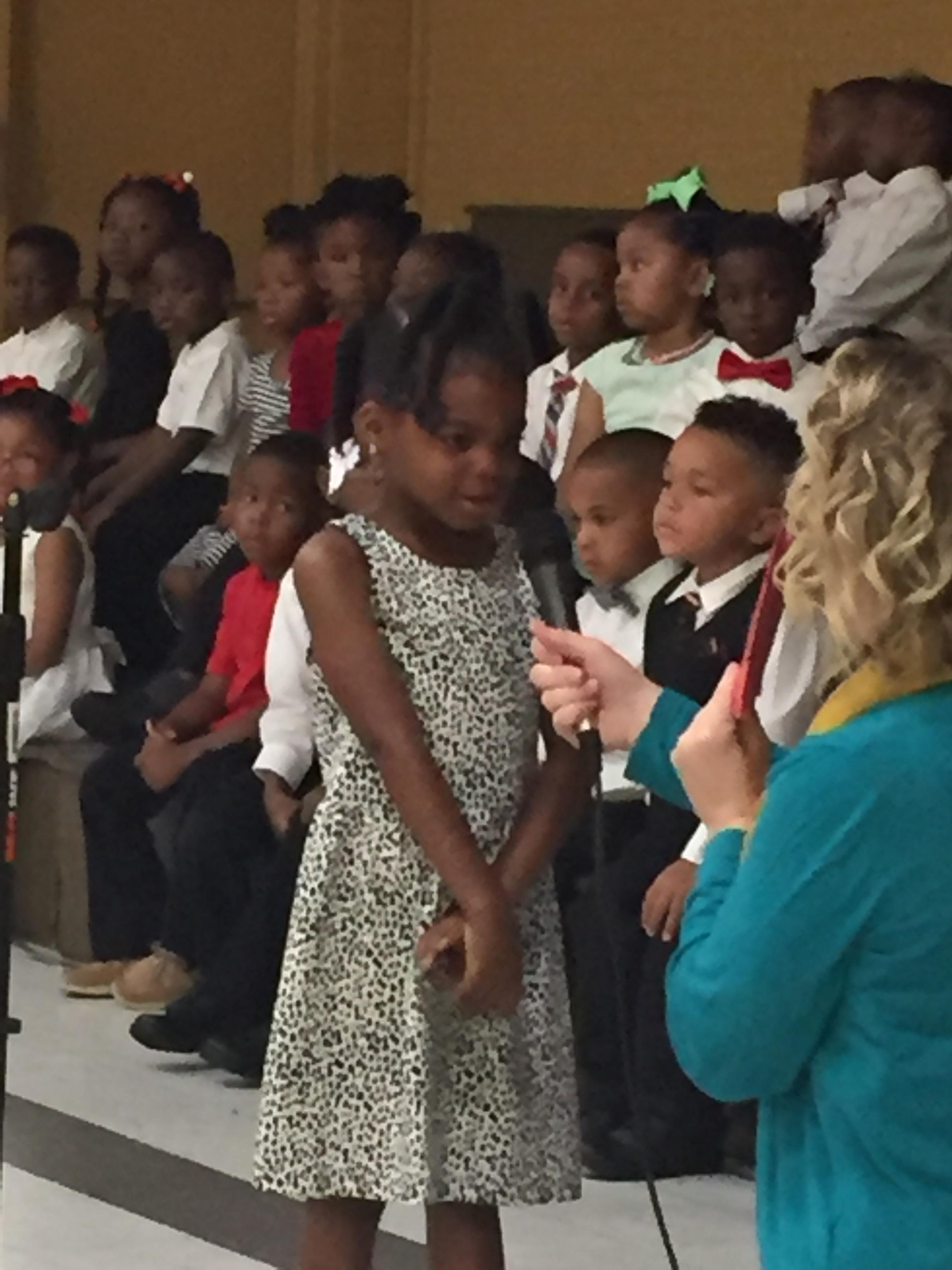 Photo of pre-kindergarten and kindergarten students at end of year awards program; a teacher is holding a microphone to the mouth of a student who is reciting
